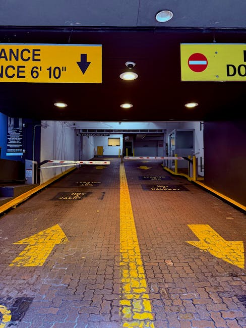 View of a vehicle entrance to a parking garage with a height restriction sign indicating a 6 feet 10 inches clearance. The ramp is paved with brick and marked with yellow directional arrows and lane lines for vehicles. On the left, there is a barrier gate that is currently closed, with yellow and black safety markings. Overhead, there are ceiling lights and signs, including a yellow sign with black text warning about the clearance height, and a red and white no-entry sign on the right. This setting appears to be part of a residential or commercial building's parking facility, suitable for vehicle access during a home relocation or furniture transport process. The image likely relates to moving logistics managed by companies like Man with Van Keston, supporting efficient load and unload activities within the parking area.