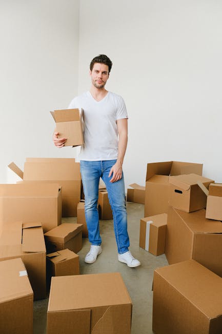A man with light skin, dark hair, and a beard stands inside a minimally decorated room with white walls and a light grey floor, holding a small cardboard box in his left hand. He is dressed in a plain white T-shirt, blue jeans, and white sneakers. Surrounding him are numerous brown cardboard boxes of various sizes, some sealed with packing tape and others open, containing packing materials such as bubble wrap and plastic wrap. The boxes are arranged both on the floor and slightly stacked, indicating an ongoing home relocation process involving packing and furniture transport. The image captures the loading or unpacking stage of a house move, with the man appearing prepared and focused on the task. This scene highlights the logistical aspects managed by companies like Man with Van Keston during moving and removals services, emphasizing the importance of careful packing and transport coordination.