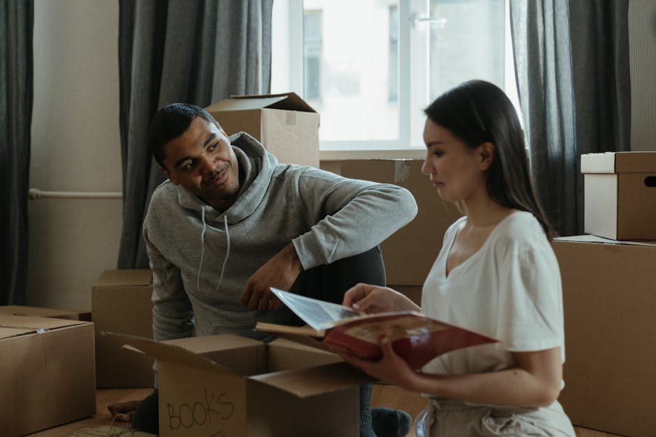 A man with dark hair tied in a bun, wearing a dark t-shirt and beige trousers, is carrying a medium-sized cardboard box with a red 'H' symbol and clear packing tape, down a staircase inside a residential property. The staircase has a wooden handrail and white walls, with a closed wooden door visible in the background. The man is holding the box firmly with both hands, preparing to transport it as part of a home relocation process facilitated by Man with Van Keston, indicated by the professional moving service setting. The lighting is bright and natural, highlighting the careful handling of household packing materials, including cardboard boxes and cushioning. This scene captures a typical furniture transport and packing phase during house removals, emphasizing careful lifting and internal logistics before external transportation begins.