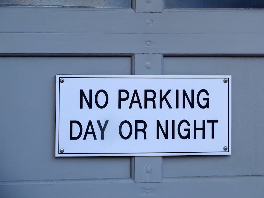 A rectangular white metal sign mounted on a grey wooden door or wall with visible panel lines and a horizontal support beam, displaying the message 'NO PARKING DAY OR NIGHT' in bold black uppercase letters. The sign is attached with four metal screws, one in each corner. The background suggests an outdoor environment, potentially on a garage door or building exterior. This sign is positioned near the top of the door or wall section. The scene is well-lit with natural lighting, highlighting the sign's clear, high-contrast text. The image reflects aspects of parking restrictions that could impact home relocation or furniture transport logistics, as part of a broader discussion on house removals, managed by a professional service like Man with Van Keston, who might need to consider street closure rules for efficient moving services near Keston and Bromley.
