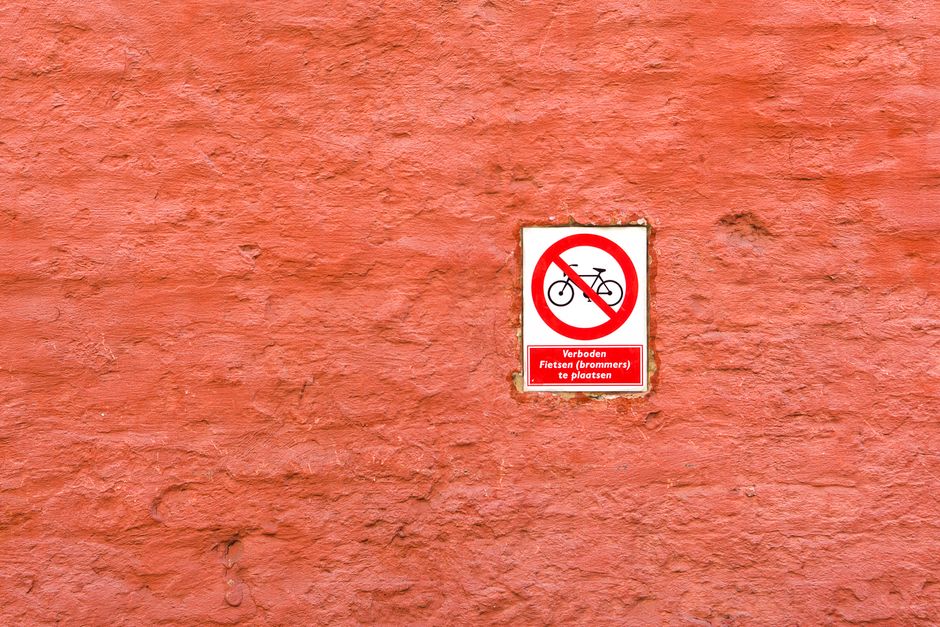 Close-up image of a red, textured brick wall with a rectangular sign mounted on it. The sign features a black bicycle symbol crossed out with a red circle and diagonal line, indicating no bicycle parking or riding allowed. Below the symbol, there is a red background section with white text in Dutch reading, 'Verboden Fietsen (brommers) te plaatsen', which translates to 'No bicycles (mopeds) to be placed'. The lighting is natural, and the sign is centrally positioned on the wall. The overall scene suggests a designated area potentially near an entrance or wall of a building where restrictions on bicycle parking are enforced, relevant to local parking regulations for home relocations or moving logistics. Man with Van Keston, a house removals service, would be familiar with such signage when planning street access or parking during moving days.