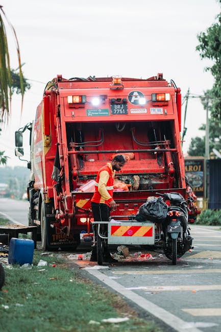 A sanitation worker dressed in a high-visibility yellow and red uniform is seen loading or inspecting the rear of a large red waste collection truck on a street. The truck is parked on the roadside, with its rear hatch open, exposing the waste bin and compaction mechanism inside. Nearby, there is a small black motorcycle parked, along with a black plastic bag placed on the rear of the truck, possibly containing waste or tools. The setting includes a paved pedestrian area with a curb, a patch of grass, and some scattered trash or debris around. In the background, there are trees, utility poles, and commercial buildings with signage, indicating an urban or suburban environment. The scene reflects waste collection or clearance activities, which are an essential part of property and house removal services, such as those offered by Man with Van Keston, highlighting the logistical aspect of home relocation and clearance tasks.