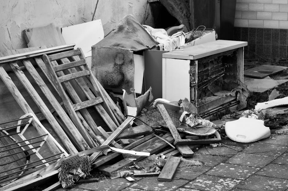 A black and white image showing a pile of discarded household items and waste materials positioned outside a residential property. The pile includes wooden items such as a dismantled bed frame or shelving unit, along with a metal wire rack, various pieces of broken furniture, and debris scattered across a paved area. There is a large, damaged appliance or piece of furniture with exposed internal components, possibly an old oven or heater. Packaging materials, including cardboard and plastic, are mixed with the debris. The scene appears to be in a cluttered outdoor space near a brick wall and a doorway, suggesting cleanup or waste removal in preparation for a house move or renovation. The overall context indicates the collection of bulky waste or furniture removal, aligned with professional removals services offered by Man with Van Keston.