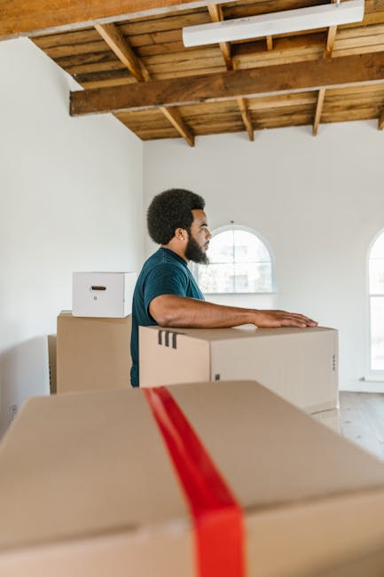 A man with dark curly hair and a beard, wearing a teal t-shirt, is standing inside a bright, spacious room with white walls and large arched windows. He is positioned behind several stacked cardboard moving boxes, some of which are sealed with packing tape, while others are open or partially obscured. The ceiling above displays exposed wooden beams and a natural wood finish, contributing to the room's open, airy atmosphere. The man appears to be preparing for a home relocation or furniture transport, with his hands resting on the boxes, possibly involved in loading or packing activities. Visible outside the windows is a blurred outdoor scene suggesting a residential area. The overall setting indicates a professional house removal process, supported by the presence of packing supplies and the focus on the careful handling of boxes, which are typical elements in moving and relocation services provided by companies like Man with Van Keston.