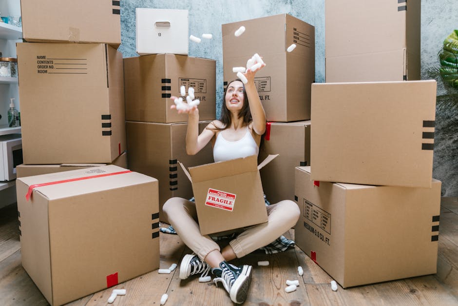 A woman sitting cross-legged on a wooden floor surrounded by numerous cardboard moving boxes, some labeled with handling instructions, inside a room with a concrete-textured blue wall and a potted plant visible on the right. She is wearing a white sleeveless top, beige pants, and black-and-white sneakers, and is smiling while tossing packing foam peanuts into the air. One box on her lap is marked with a red 'Fragile' sticker. The boxes vary in size, with some stacked on top of each other, and others placed on the floor, indicating an ongoing packing or moving process. Visible equipment includes the foam peanuts she is handling, and the scene likely depicts a home relocation or furniture transport activity, with [COMPANY_NAME] providing professional removals in preparation for a house move in Keston.