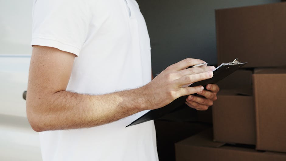 Close-up of a person wearing a white t-shirt, holding a black clipboard and writing with a pen. The individual is standing indoors, likely in a room designated for packing or storage during house removals. Behind them, there are stacked cardboard boxes, some wrapped in plastic or fabric covers, indicating packing materials used in a home relocation process. The scene captures part of their arm and torso, with focus on the clipboard and the act of writing, suggesting they are documenting inventory or planning logistics for a furniture transport or moving service. The environment appears well-lit with natural or indoor lighting, and the setting aligns with professional removals activities carried out by companies such as Man with Van Keston, supporting efficient packing and moving operations.