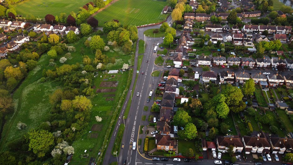 An aerial view of a residential street showing rows of houses with varied rooftops, surrounded by lush green trees and gardens. The street features parked cars along the roadside and a central lane with marked vehicle restrictions, indicating a quiet neighbourhood. To the left side of the image, there is a large garden area with neatly cultivated vegetable plots, bordered by a line of mature deciduous trees with fresh spring foliage. The scene is well-lit with natural daylight, illustrating a local community suitable for home relocations and furniture transport logistics. Man with Van Keston, a professional removals service provider, operates within this type of environment, assisting with packing, loading, and moving processes required for house removals in the Keston area.