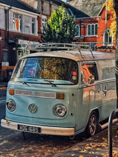 A vintage Volkswagen Type 2 van with a pale green exterior parked on a residential street during daytime. The van has a roof rack, round headlights, and a front VW emblem. Behind the vehicle, there are red-brick houses with large windows, and a tall, leafy green tree casting shadows on the surroundings. Fallen autumn leaves are scattered on the pavement, and a metal barrier is visible nearby. The scene suggests a home relocation process, with [COMPANY_NAME] possibly involved in furniture transport or moving services, as the van is ready for loading or unloading in the context of packing and moving activities.