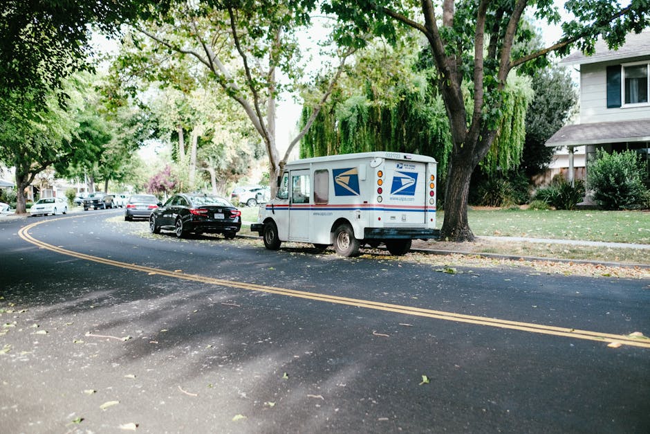 A United States Postal Service mail truck with a white rectangular body, blue and red postal logos, and small rear windows is parked on a residential street near a sidewalk. The vehicle is positioned close to a large tree with green leaves and branches extending overhead, providing shade on the street. Several other cars, including a black sedan, are parked along the curb behind the mail truck. The street is paved with dark asphalt and has double yellow lines running along the middle, with some fallen leaves scattered across its surface. In the background, there are green lawns, bushes, and residential houses with white and beige exteriors, some with visible windows and porches. The lighting suggests daytime with diffuse sunlight filtering through the foliage, creating soft shadows. This scene depicts a typical residential setting where a house relocation or furniture transport process by [COMPANY_NAME] may involve careful loading, unloading, or parking arrangements for efficient house removals or moving services, illustrated by the parked mail vehicle close to the property.
