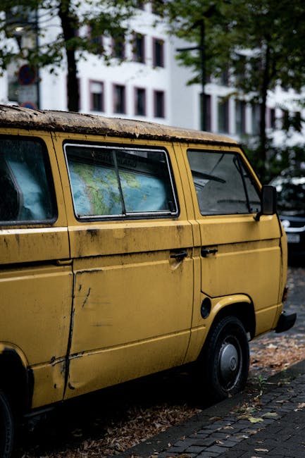 A vintage Volkswagen Type 2 van with a pale green exterior parked on a residential street during daytime. The van has a roof rack, round headlights, and a front VW emblem. Behind the vehicle, there are red-brick houses with large windows, and a tall, leafy green tree casting shadows on the surroundings. Fallen autumn leaves are scattered on the pavement, and a metal barrier is visible nearby. The scene suggests a home relocation process, with [COMPANY_NAME] possibly involved in furniture transport or moving services, as the van is ready for loading or unloading in the context of packing and moving activities.