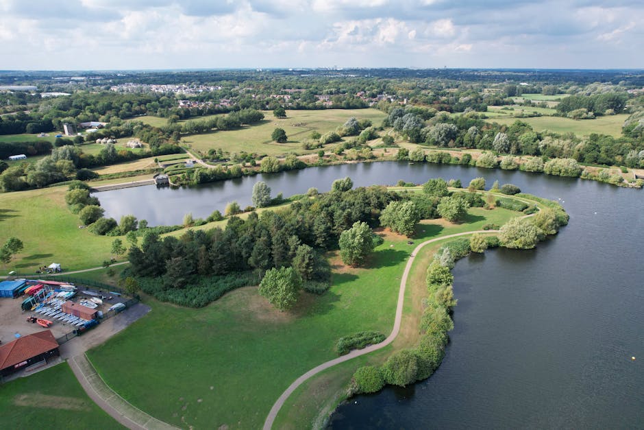 An aerial view of Keston Park Estate with a large body of water surrounded by green lawns, trees, and landscaped gardens. A winding footpath runs along the edge of the water, leading to a small wooded