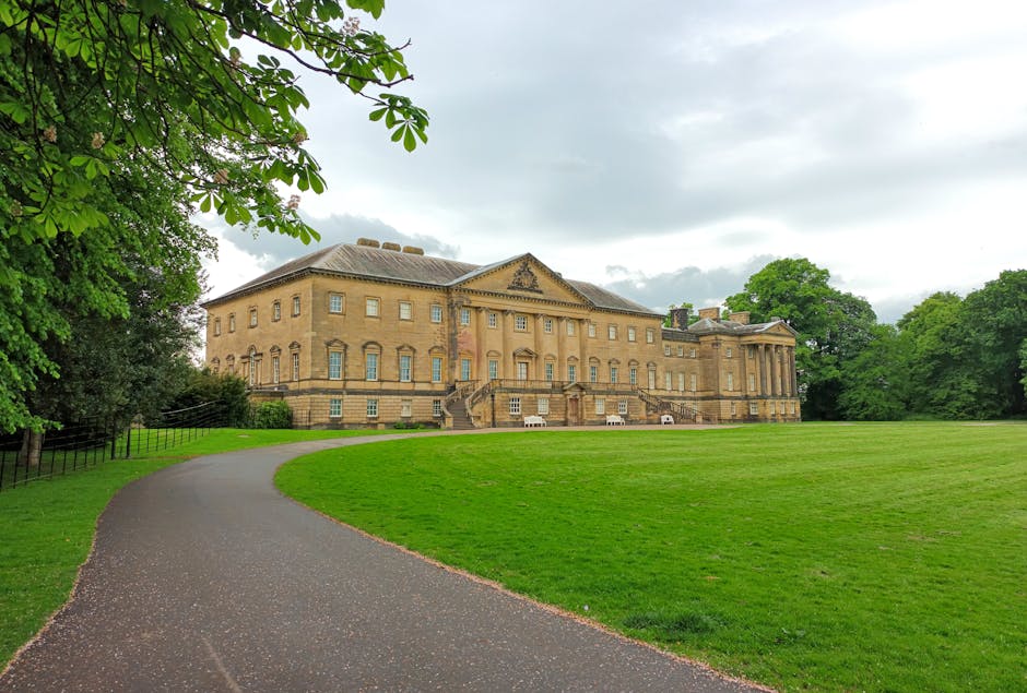 An aerial view of Keston Park Estate with a large body of water surrounded by green lawns, trees, and landscaped gardens. A winding footpath runs along the edge of the water, leading to a small wooded