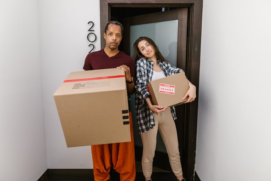 A man and a woman standing inside a residential building near a doorway, both holding cardboard moving boxes. The man, dressed in an orange pair of pants and a burgundy shirt, is holding a large box with red and black tape, while the woman, wearing beige trousers and a casual plaid shirt over a white top, carries a smaller box marked with a red 'FRAGILE' label. The environment is well-lit with natural or artificial lighting, and the doorway appears to lead to the outside or a corridor. The scene illustrates the process of home relocation, with the individuals preparing to load the boxes into a vehicle or transport them within the property. This visual aligns with house removals and local moving services offered by Man with Van Keston, highlighting packing, handling, and the logistical aspects of furniture transport and moving preparations.