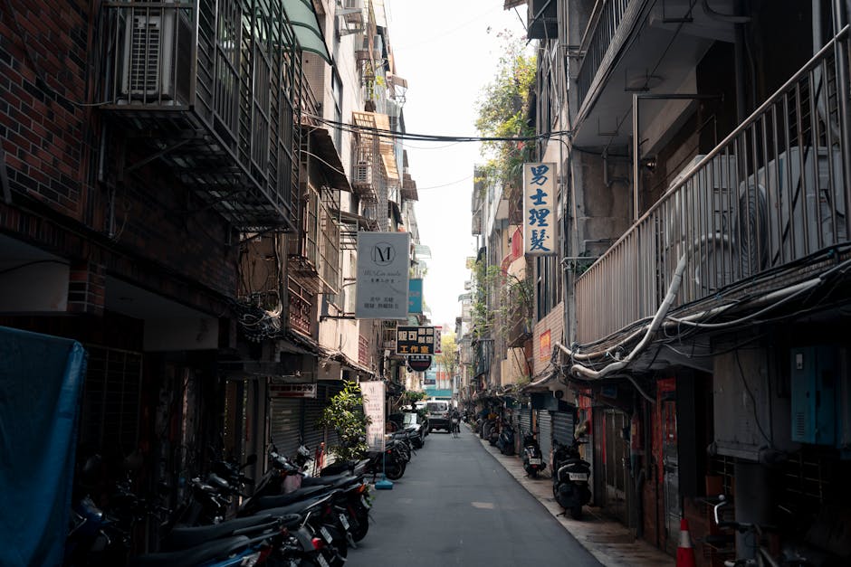 A narrow urban street with tightly packed multi-storey buildings on both sides, featuring metal fire escape stairs and balconies adorned with various signs, including Asian script and English text. The street is lined with parked scooters and motorcycles, while a delivery vehicle is visible further down the road. Overhead, an array of electrical wires crisscross above the street. The scene is illuminated by natural daylight, highlighting the confined space. This setting exemplifies the challenge of navigating tight access areas during home relocation or furniture transport, where professional removals from Man with Van Keston are often required to carefully manage loading and unloading processes within limited space constraints.