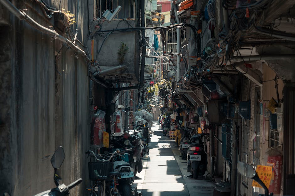 A narrow urban street with tightly packed multi-storey buildings on both sides, featuring metal fire escape stairs and balconies adorned with various signs, including Asian script and English text. The street is lined with parked scooters and motorcycles, while a delivery vehicle is visible further down the road. Overhead, an array of electrical wires crisscross above the street. The scene is illuminated by natural daylight, highlighting the confined space. This setting exemplifies the challenge of navigating tight access areas during home relocation or furniture transport, where professional removals from Man with Van Keston are often required to carefully manage loading and unloading processes within limited space constraints.