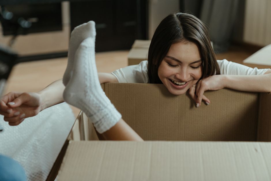 A young woman with shoulder-length brown hair, smiling with closed eyes, leans over a large cardboard moving box inside a room. She is wearing a white t-shirt and white socks, with her legs extended u