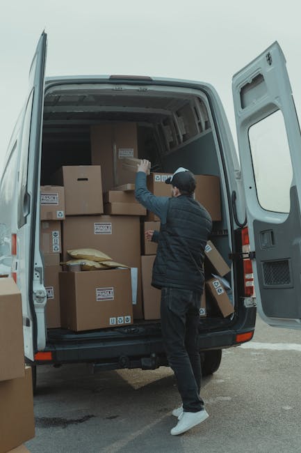 A man with a beard, wearing a black cap, white polo shirt, and blue jeans, stands inside the open back of a large white moving van. He is holding a cardboard box with both hands, positioned in front o