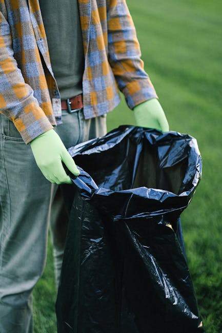 An outdoor view of a large, overfilled waste disposal area containing multiple black, green, and red trash bins, with some bins overflowing and cardboard boxes and plastic bags spilling out onto the s